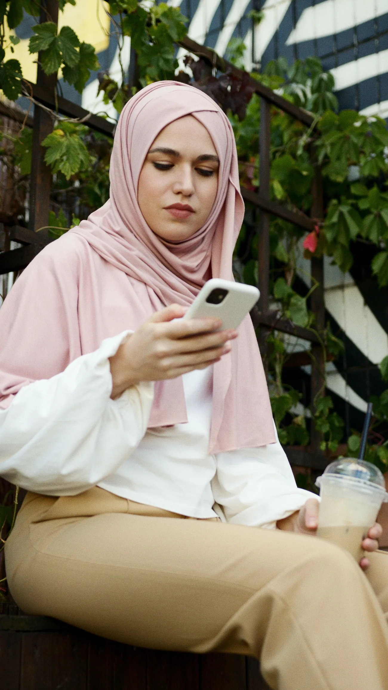 A young woman in a pink headscarf smiling at her phone as she types a message