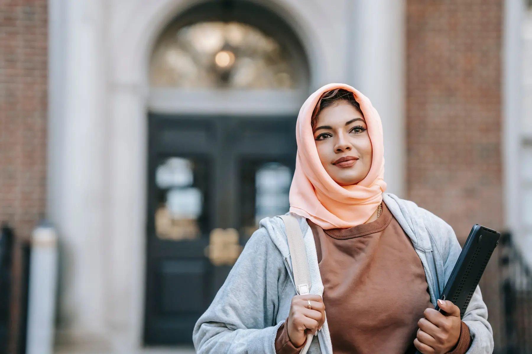 A smiling student in a headscarf holding a folder outside a university doorway