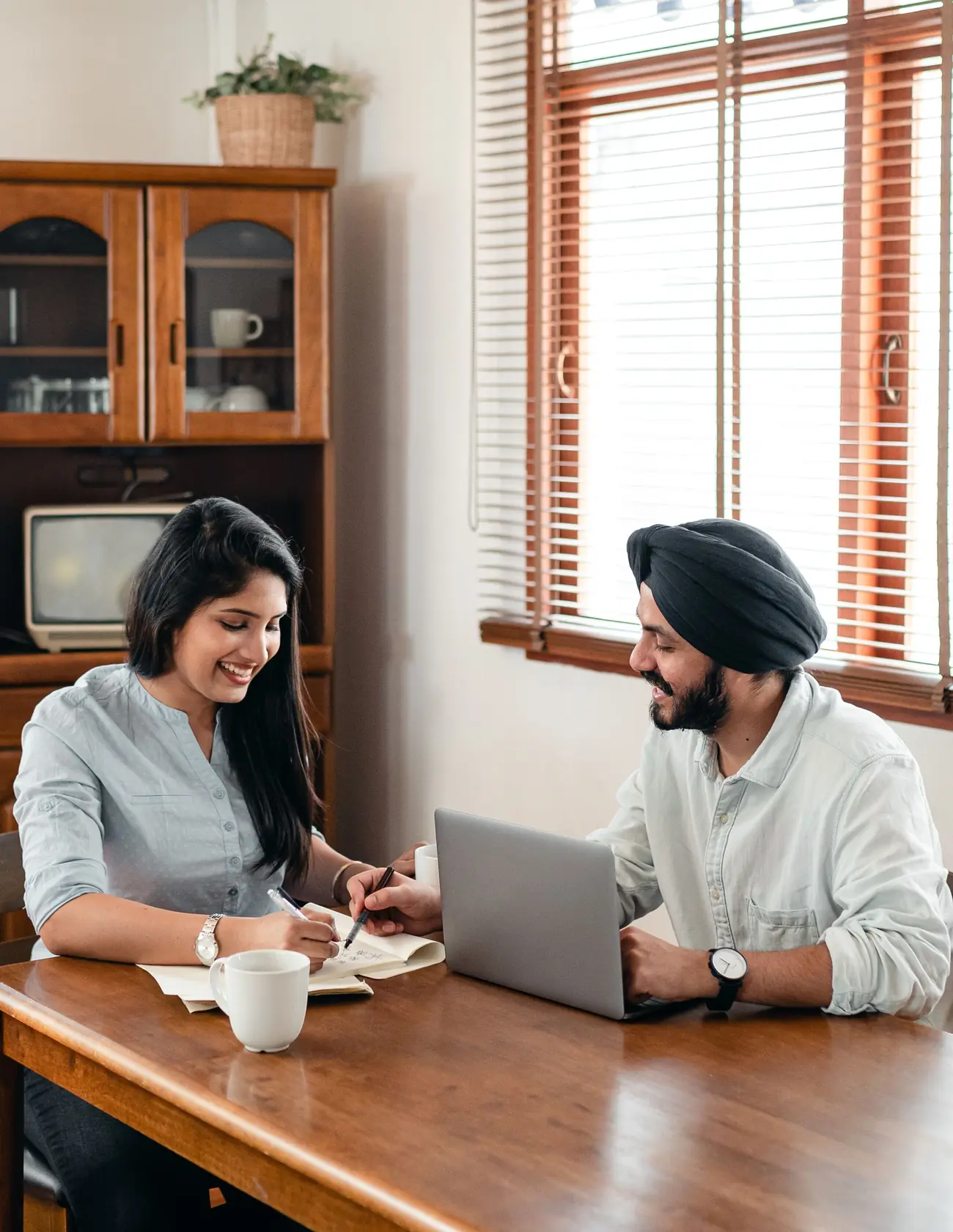 Two students laughing over homework at a kitchen table with a laptop