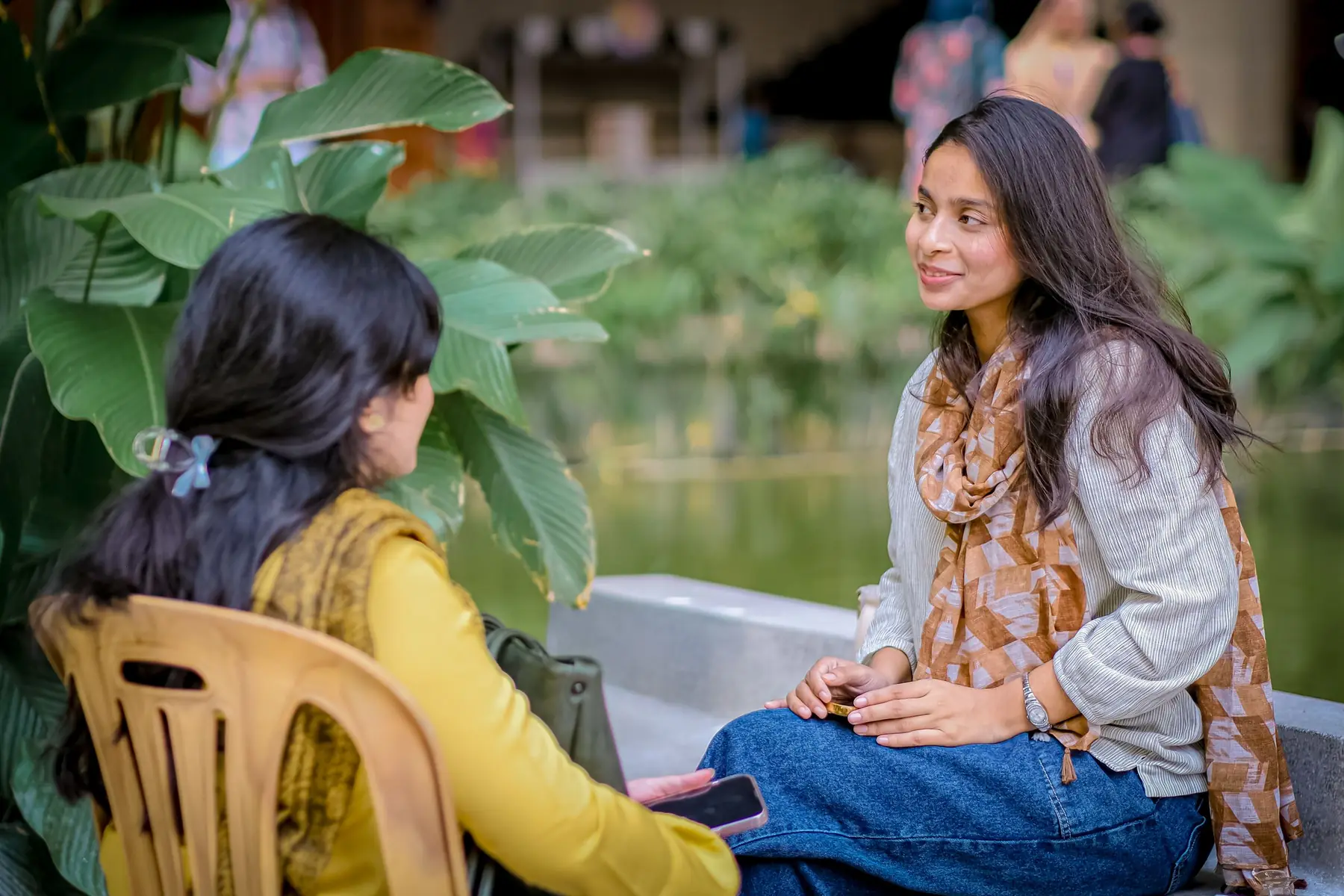Two young women in conversation on a bench beside a garden pond